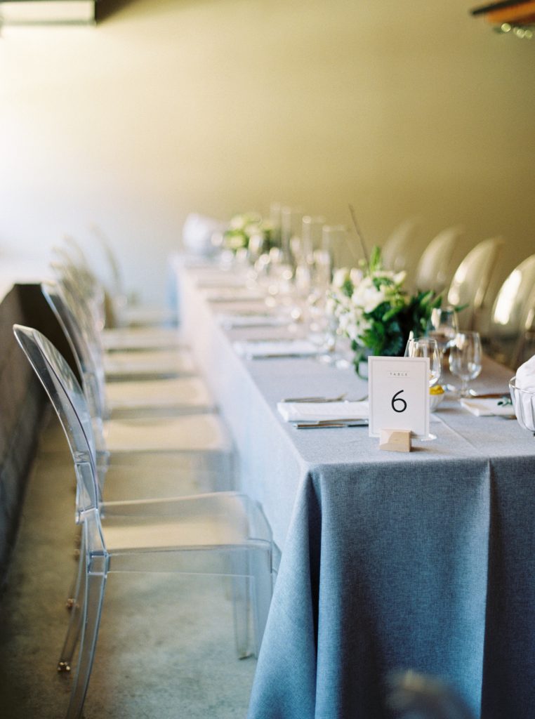Minted table numbers, grey tapered candles on grey wedding linen with ghost chairs and loose green and white floral arrangements by Blooms and Flora at Alton Mill Wedding | Goldie Mill Wedding | Caledon Wedding Photographer | Guelph Wedding Photographer | Kayla Yestal