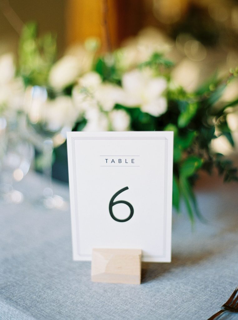 Minted table numbers, grey tapered candles on grey wedding linen with ghost chairs and loose green and white floral arrangements by Blooms and Flora at Alton Mill Wedding | Goldie Mill Wedding | Caledon Wedding Photographer | Guelph Wedding Photographer | Kayla Yestal